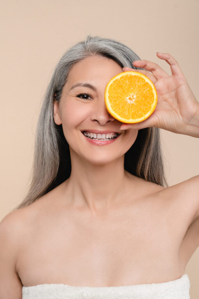middle aged woman posing wearing a towel, smiling and holding half an orange covering half her face